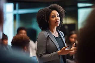A professional Black woman with curly hair presenting to colleagues in a meeting room, gesturing with her hands as she speaks