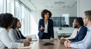 A woman in a navy blazer standing and presenting to a diverse group of colleagues around a conference table in a modern glass-walled office