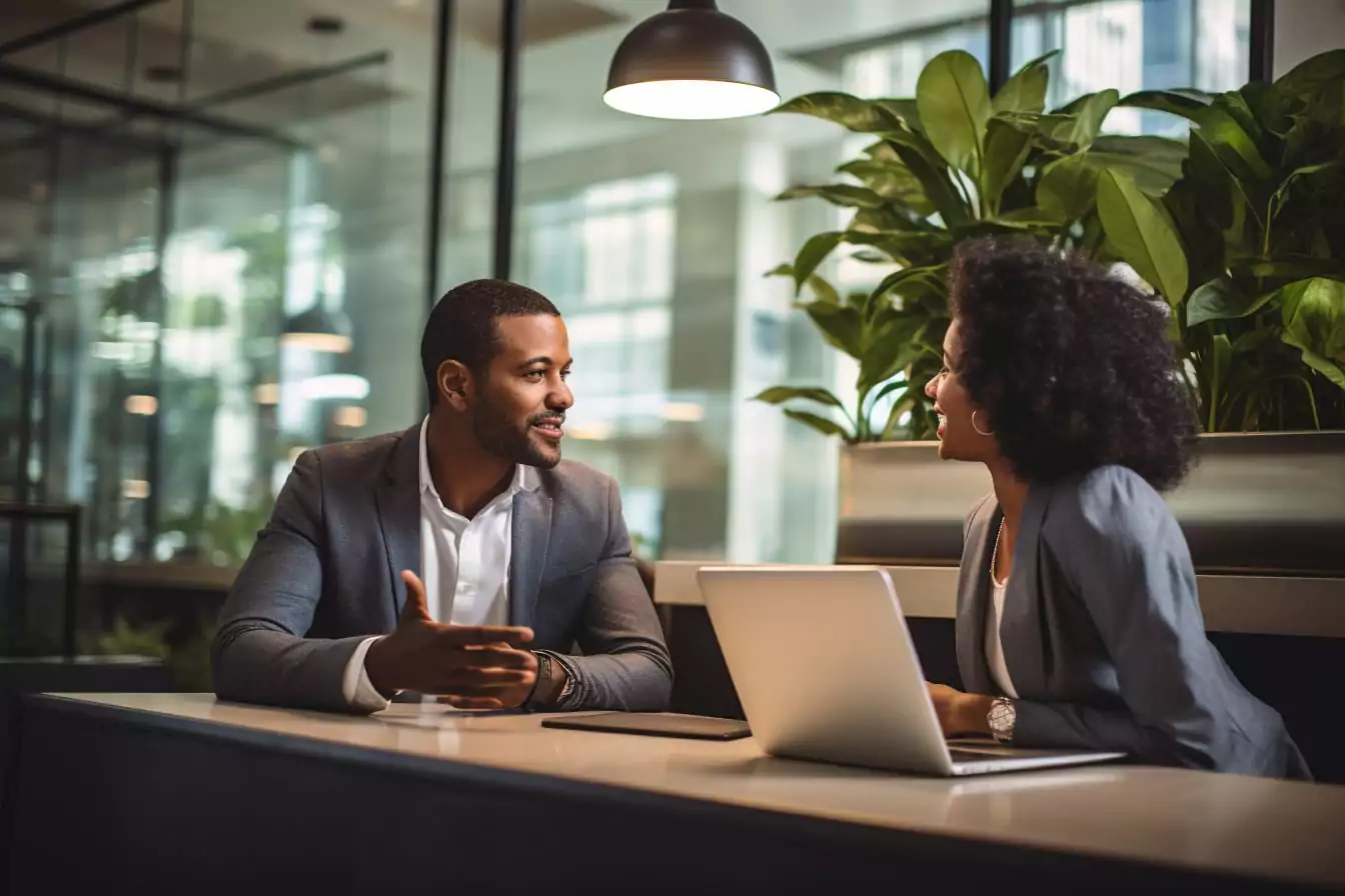 A Black man in a gray suit and a Black woman with curly hair having a business conversation over a laptop in a well-lit office space.