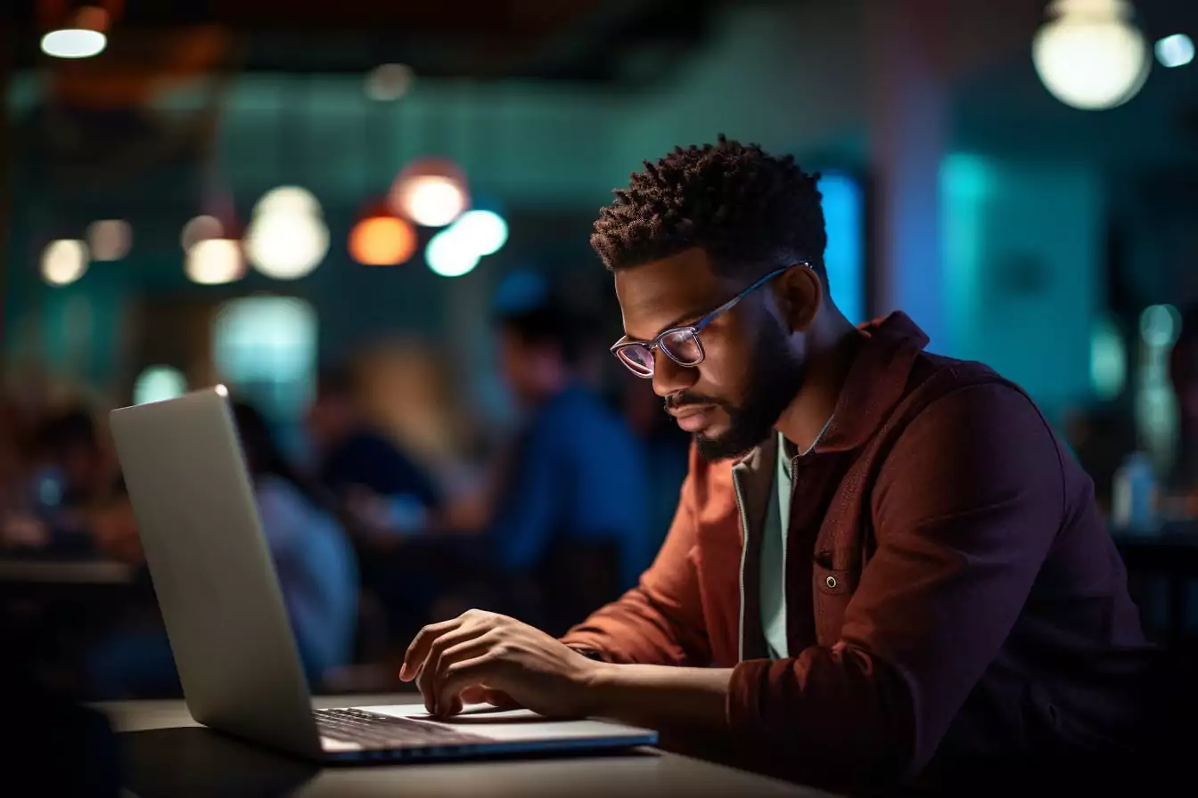 Black man with glasses and a beard concentrating on his laptop in a dimly lit room with a blue ambiance, indicating evening work.